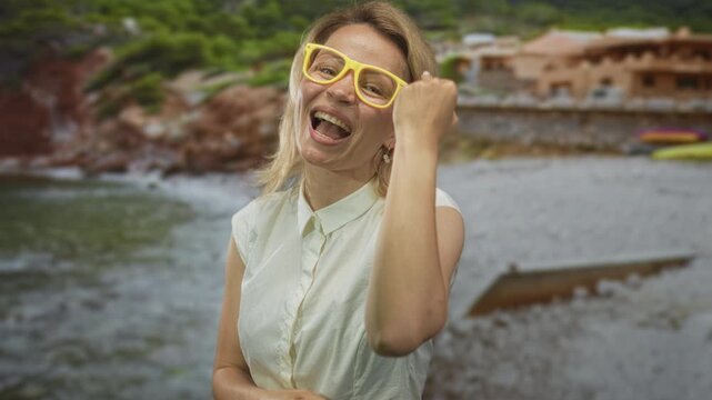 Woman wearing yellow glasses shows middlefinger at sunlit building by seaside; raw rebellious defiance.