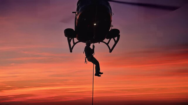 A person is rappelling down from a helicopter against a vibrant orange and purple sunset sky