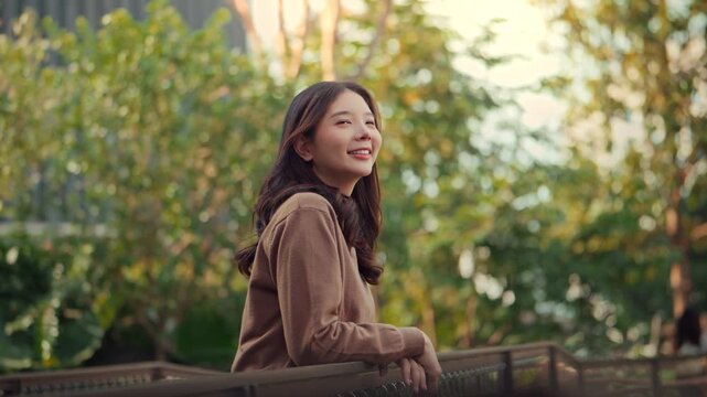 Happy young Asian woman leaning on a balcony railing and looking away with a smile in a lush green park. Concept of mental health wellness, relaxation, and mindfulness during a peaceful 