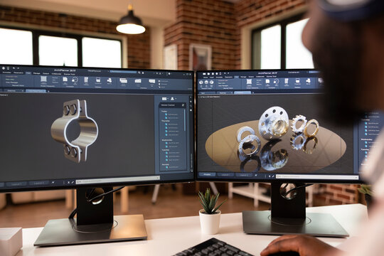 African american developer working in home studio with dual monitors and CAD tools. Male engineer develops metal components with 3D modeling software, visualizing industrial designs with precision.