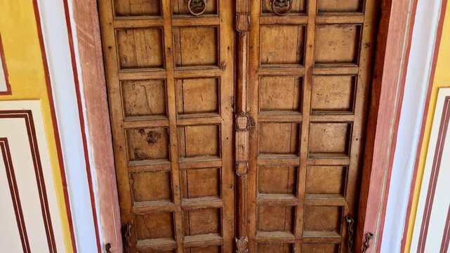 An antique wooden door with intricate paneling and circular brass knockers stands within the Kanak Vrindavan Mahal. Its weathered texture and traditional latch reflect the historic charm of Jaipur.