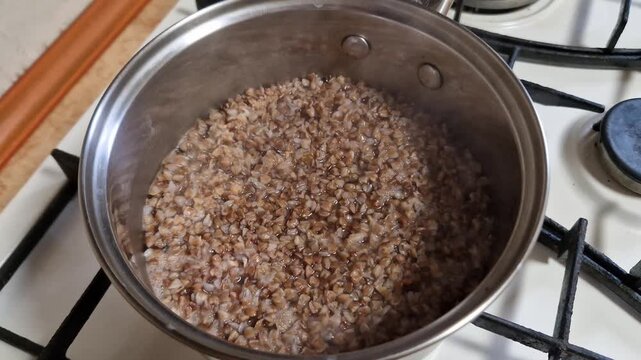 Close-up overhead shot of boiling buckwheat grains in a stainless steel pot on a white gas stove with gentle steam rising. Healthy food preparation for a meal in a domestic kitchen setting.