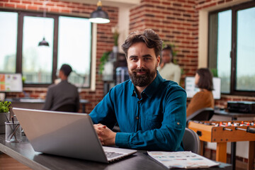 Fototapeta premium Caucasian male researcher seated at desk with crossed hands, gazing at camera in modern workspace. Portrait of young manager with laptop at table for research and reviewing business project plans.