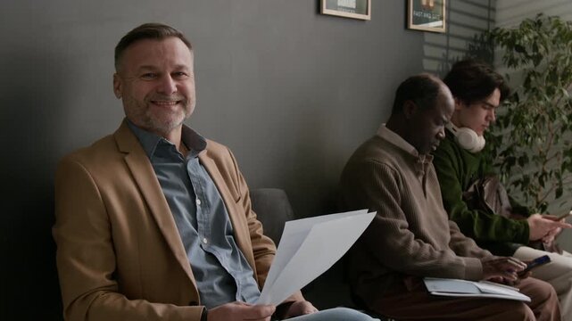 Portrait shot of positive middle aged man in blazer smiling while sitting with documents in waiting area of modern bank