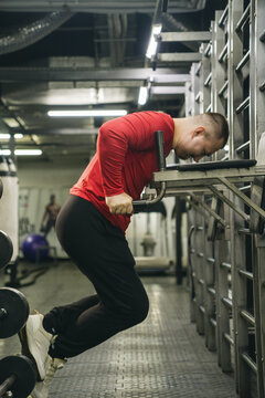 A man in a bright red compression shirt and black pants executes a dip exercise on parallel bars in a dimly lit, industrial-style gym setting, focusing intensely on the workout.