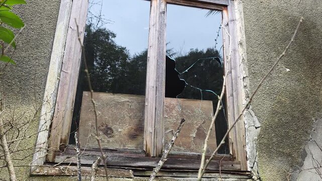Broken window glass of an abandoned building reflecting the surrounding nature and cloudy sky