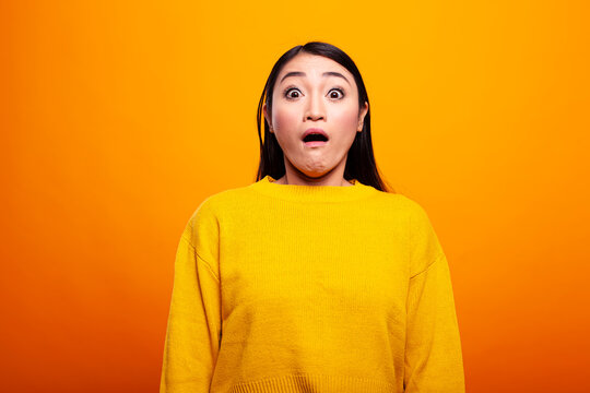 Young woman in a yellow sweater shows a surprised and fearful expression, with wide eyes, staring in awe. Asian female adult looking at camera with her mouth open, appearing shocked and stunned.