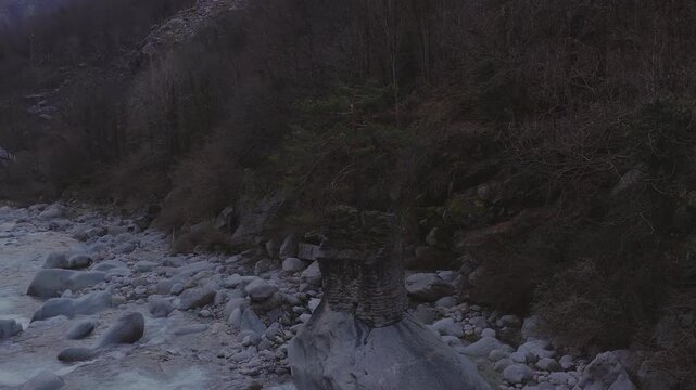 Aerial shot shows a pale turquoise mountain stream in Italy, a ruined stone bridge pier with a pine, rounded boulders, and leafless slopes in cool low light.