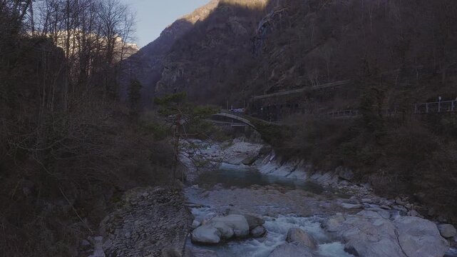Aerial view shows a stone arched bridge in Italy as a small blue clad person stands, a turquoise river ripples around rocks, steep leafless slopes and sunlit peaks frame scene