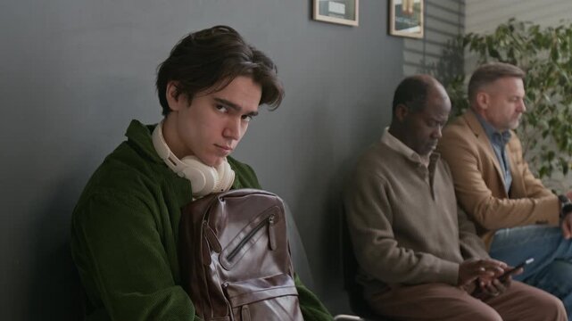 Tilt up portrait shot of young man in green hoodie, wearing headphones and carrying backpack, looking at camera, while two men sitting behind him in bank waiting area