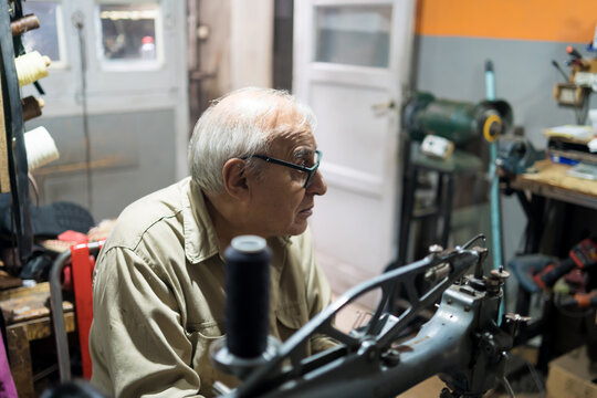 Elderly Latino upholsterer focused on repairing a backpack with a sewing machine