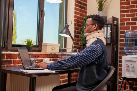 Black businessman with cervical collar typing on personal computer at brick wall office, concentrating on project development after accident. Injured male employee analyzing strategy ideas on laptop.