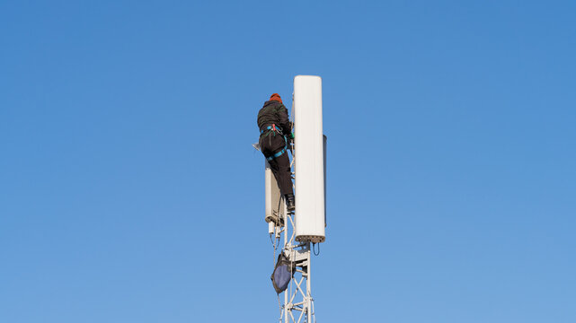 Worker climber installing a cell phone tower antenna. An industrial climber technician installing a new cellular antenna unit on a single pole telecommunications mast. Telecom infrastructure services.