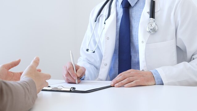 Adult aged doctor man taking notes while listening to a female patient explaining her symptoms during a medical consultation in the clinic office. Medicine and health care concept