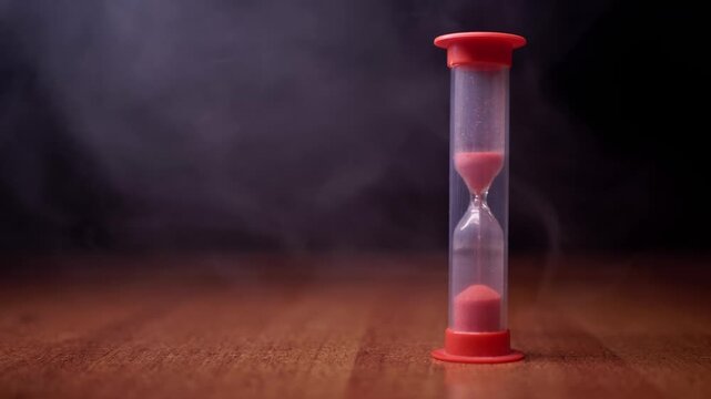 A red hourglass with pink sand sits on a wooden table as smoke drifts in the dark background. Concept of deadline pressure, time running out, and the fleeting nature of moments.