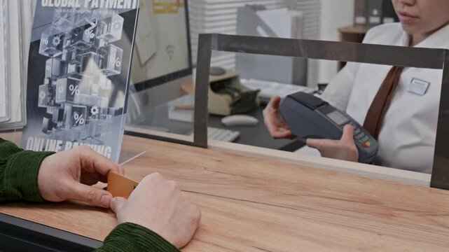 Close up shot of unrecognizable man paying with credit card, standing at wooden counter with galss wall in bank office