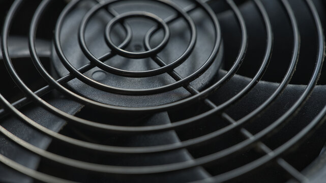 A dusty black computer power supply unit fan with a circular metal grill and visible dust particles. Shallow depth of field. Close up.