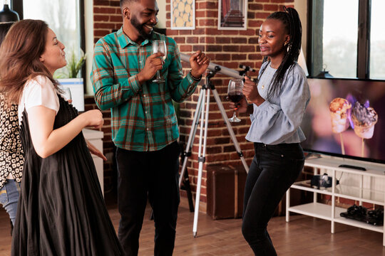 Cheerful african american couple, happily dancing, drinking wine, alcohol accompanied by caucasian young adult female friend. Partners enjoying friendship, partnership, social life.