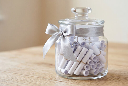 Glass jar with rolled white paper notes tied silver bow on table