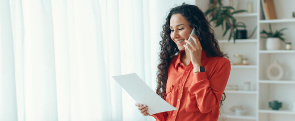A woman stands in a bright room holding a piece of paper while talking on a phone. She appears engaged in her conversation and smiles. Plants decorate the background. © Prostock-studio