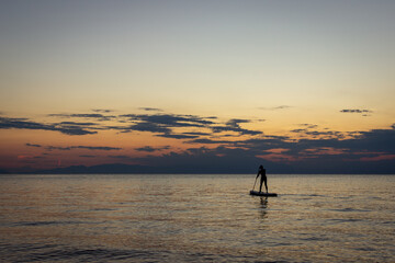 Lone paddleboarder silhouette stands on still water at dusk, framed by an orange-to-blue gradient sky and distant mountain silhouette. Evokes freedom, solitude, and outdoor adventure. © Katarina