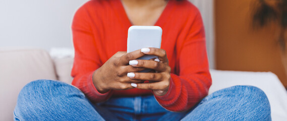Fototapeta premium A woman sits with her legs crossed on a sofa, focused on her smartphone. She wears a red sweater and jeans. The background shows a cozy living room with plants.