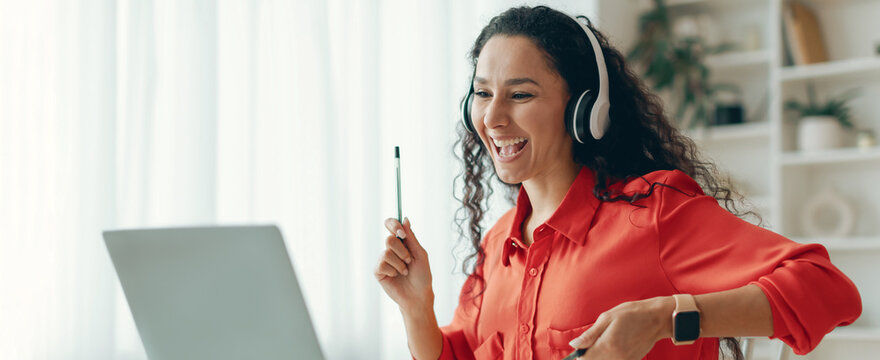 A woman is enjoying an online class or meeting while sitting at a desk. She is wearing headphones and smiling. She holds a pen and interacts with her laptop. Sunlight comes through the window.