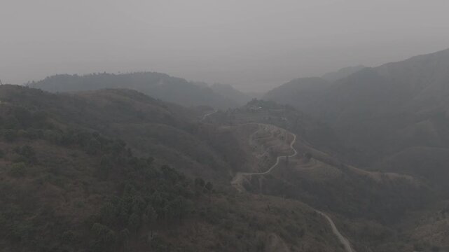 Aerial view of Dwara village near Dehradun, Uttarakhand, with a dirt path along a mountain ridge, misty hills, terrace farms, sal and pine forests, and the Song River below on a winter evening.
