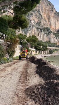 Coastal Cleanup: An industrial machine diligently clears debris along a picturesque coastal road, set against a backdrop of a majestic cliff face and lush vegetation.