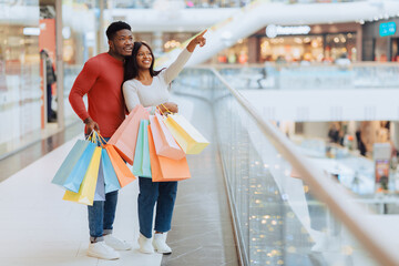 Obraz premium A man and woman are standing in a shopping mall. They hold colorful shopping bags. The man points toward something in the distance while the woman smiles.