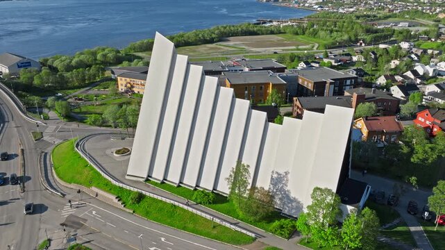 Drone aerial approaching the Arctic Cathedral in Troms&oslash;, Norway. Modern triangular church stands beside the road with fjord waters and city skyline in the background.