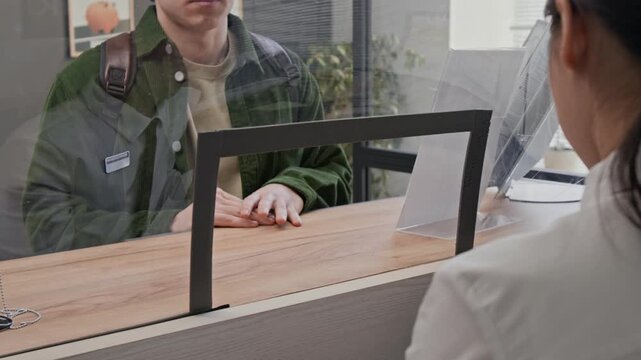 Low angle close up shot of bank teller accepting document with red stamp from young male customer, separated by transparent protective screen in bank