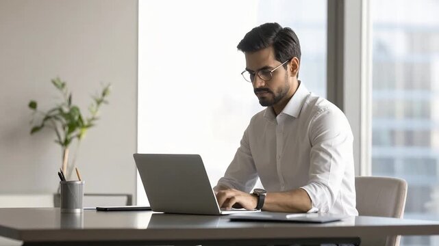 Adult man working on laptop at modern office desk near window showing focused male professional in calm indoor workspace environment