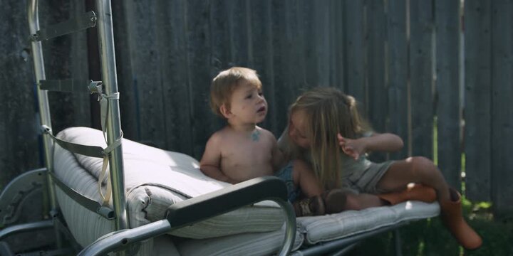 Two children sitting on a sun lounger in a field by a fence