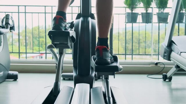 Man exercising on an elliptical trainer in modern gym with close up on legs and pedals. Male doing cardio training on elliptical machine in fitness studio. Fitness, wellness, healthy lifestyle concept