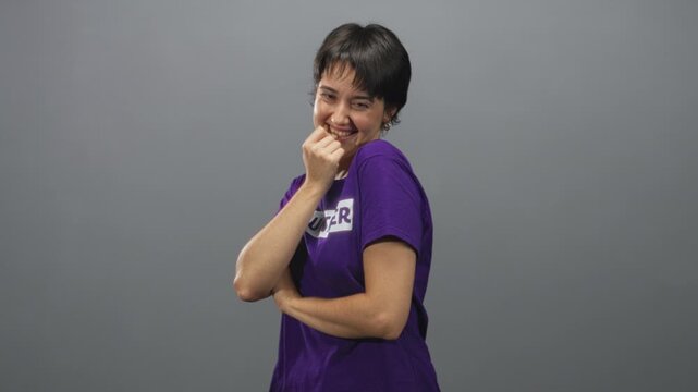 Woman biting knuckle with hand to mouth in studio wearing purple volunteer tshirt, arm folded across torso; playful shy warmth.