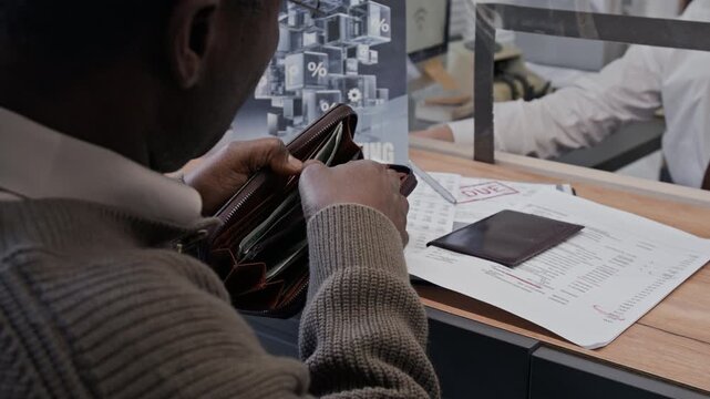 Over the shoulder shot of senior African American man giving money to young female bank teller through glass counter