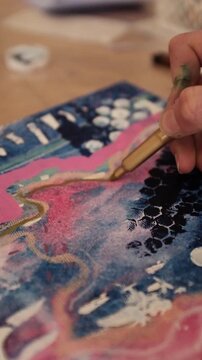 Close-up of a female artist's hands using a decorative stencil and paintbrush to apply white acrylic paint onto a textured, warm-toned abstract mixed-media canvas.