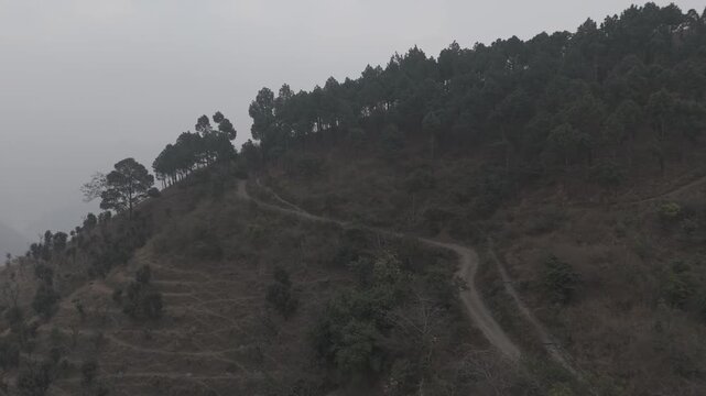 Aerial view of Dwara village near Dehradun, Uttarakhand, with a dirt path along a mountain ridge, misty hills, terrace farms, sal and pine forests, and the Song River below on a winter evening.