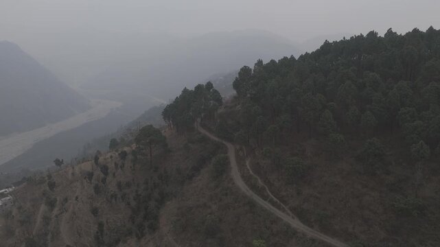 Aerial view of Dwara village near Dehradun, Uttarakhand, with a dirt path along a mountain ridge, misty hills, terrace farms, sal and pine forests, and the Song River below on a winter evening.