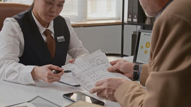 Female bank employee pointing with pen at document held by man sitting across table in bank office