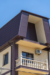 Fototapeta premium Architectural detail of modern building with dark brown mansard roof, dormer window, light yellow facade, elegant balcony with classic balustrade, and clear blue sky.