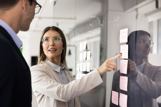 Young businesswoman engaged in discussion with male colleague while pointing at glass wall covered in colorful sticky notes, explaining strategy or insights, mapping out tasks. Brainstorming, planning