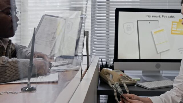Senior African American man paying money over glass counter to young female bank teller in modern bank