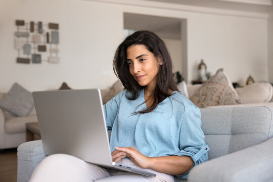 Focused young woman reviewing products or making purchases online using laptop seated on cozy sofa in living room, checking bank accounts, managing freelance assignments, scrolling social media news