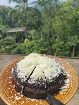 Kue cokelat lembut dengan topping keju parut melimpah di atasnya, menciptakan perpaduan rasa manis dan gurih yang menggoda. Teksturnya tampak moist dan padat, disajikan di atas piring dengan latar pem