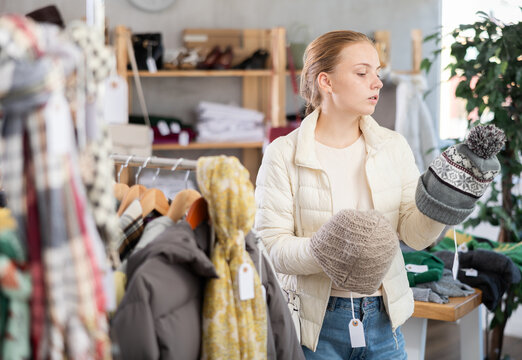 Hesitant teenage girl reviewing winter hats during sale in clothing shop