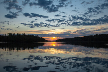 Fototapeta premium Dramatic summer sunset over Lake Ladoga with colorful clouds reflecting in the calm water. Dark silhouettes of forested shores frame the glowing horizon.