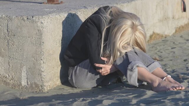 Upset blonde woman sitting barefoot on the sand, hiding her face in her hands. A portrait of helplessness, loneliness and deep sorrow