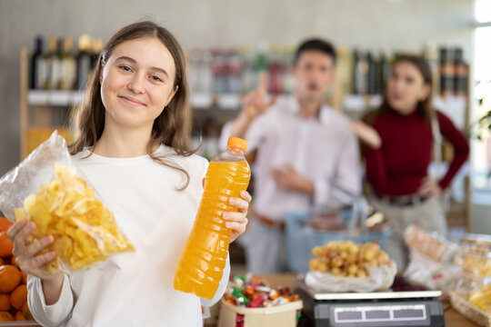 Happy teenage girl holding potato chips and juice while parents reacting discontentedly behind in grocery